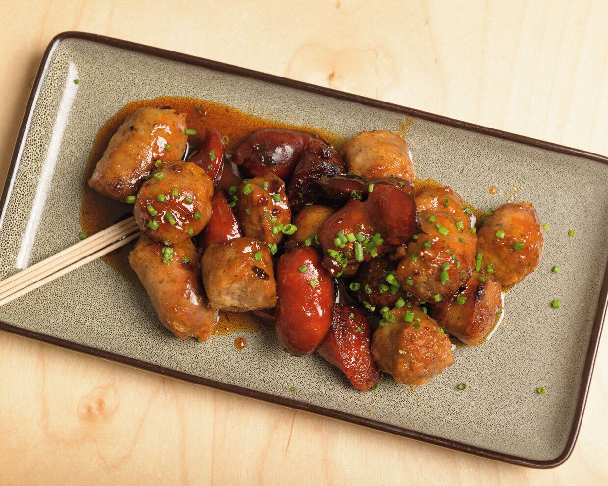 A dish of glazed meatballs and sausages garnished with chopped green onions, served on a speckled ceramic plate.