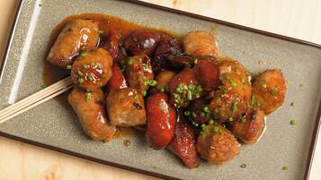 A dish of glazed meatballs and sausages garnished with chopped green onions, served on a speckled ceramic plate.