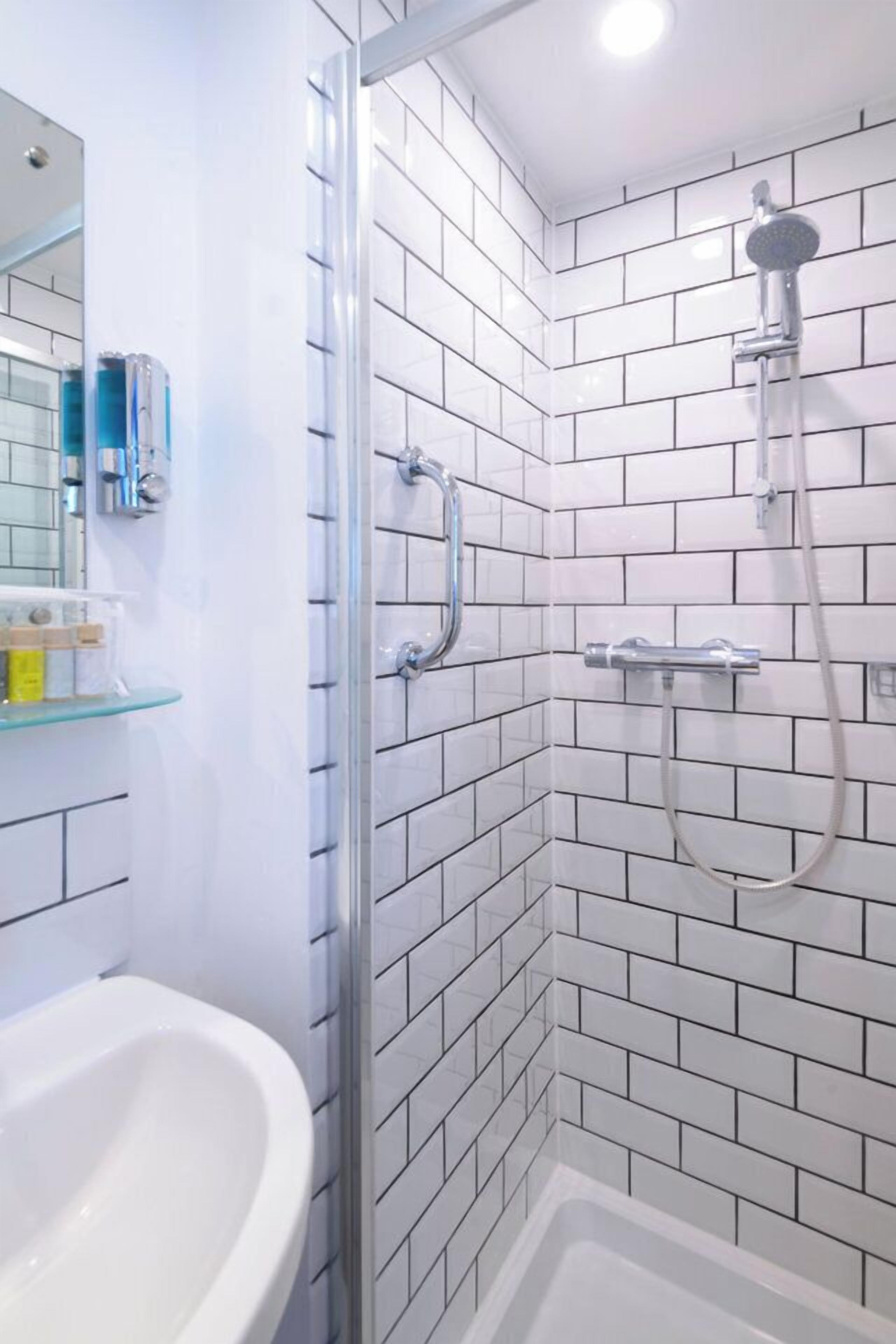 Une salle de bain moderne avec des carreaux de céramique blancs rectangulaires, une douche en verre, un lavabo blanc et une étagère en verre où sont rangés les articles de toilette.