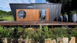 Un bâtiment moderne et compact, doté d’une terrasse en bois, d’une fenêtre circulaire et d’une porte.