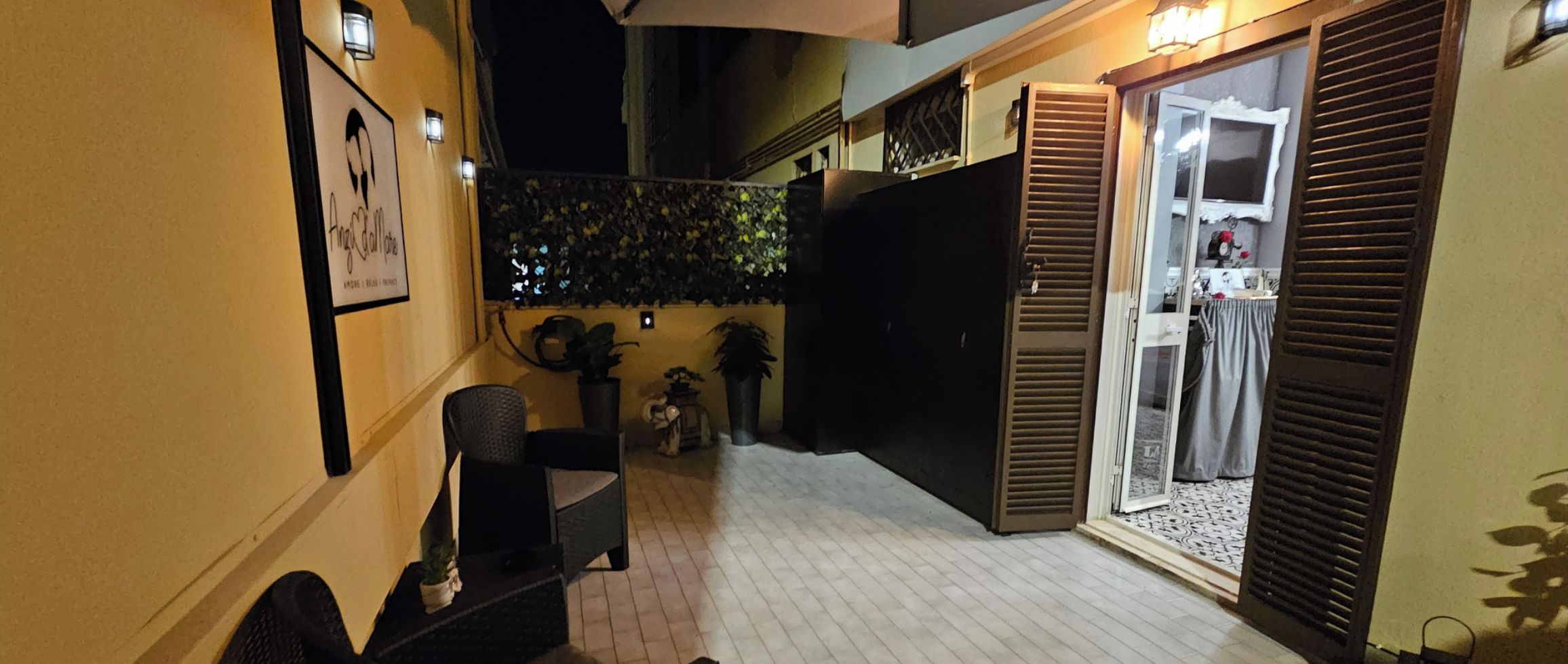 Un stretto corridoio esterno con una sedia, un tavolino, una pianta in vaso e una porta che dà su una stanza con finestra e tenda.