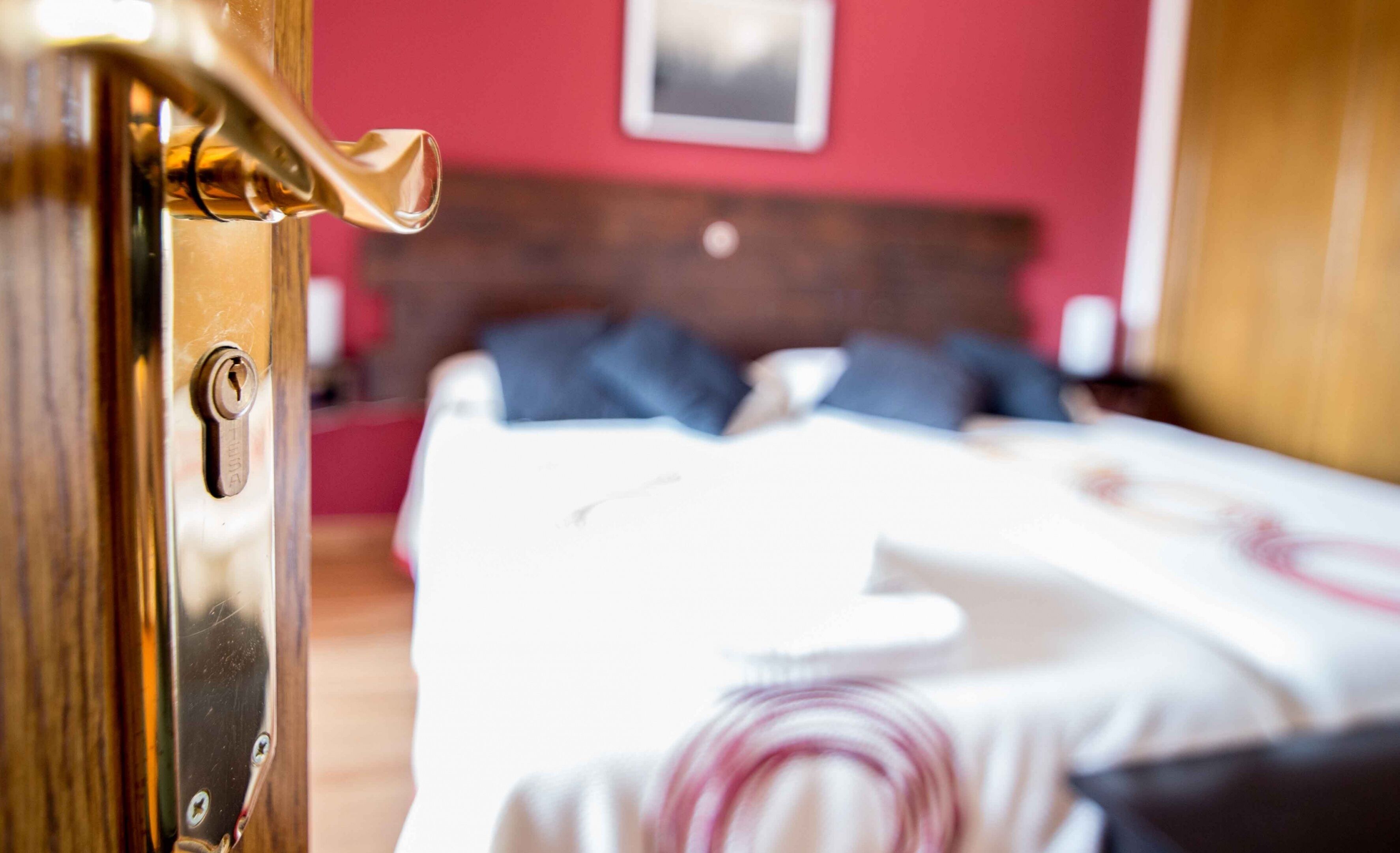 Une chambre d’hôtel avec une porte en bois, un lit recouvert d’une courtepointe à motifs blancs et roses, et un mur rouge orné d’un tableau encadré.