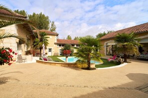A courtyard with a swimming pool, palm trees, and a building with a tiled roof. - Elegant Périgord: 'Le Pré Fleuri' Guest Room with Pool in the Heart of Ribéracois (Saint-Sulpice-de-Roumagnac)