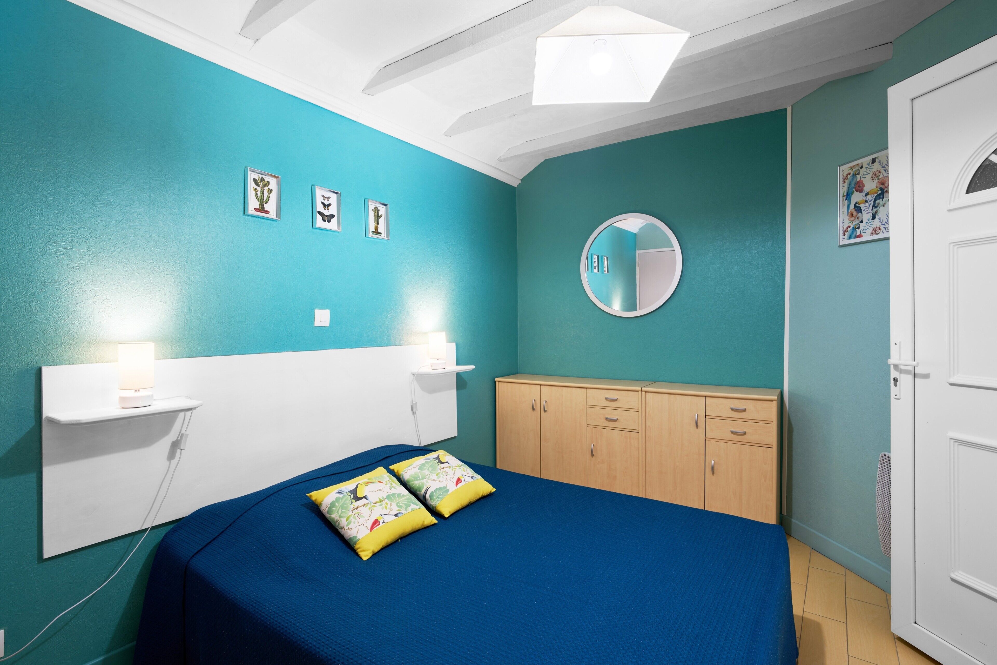 Een slaapkamer met blauwgroene wanden, een bed met blauw beddengoed, een nachtkastje met een lamp, een kledingkast en een ronde spiegel aan de muur.