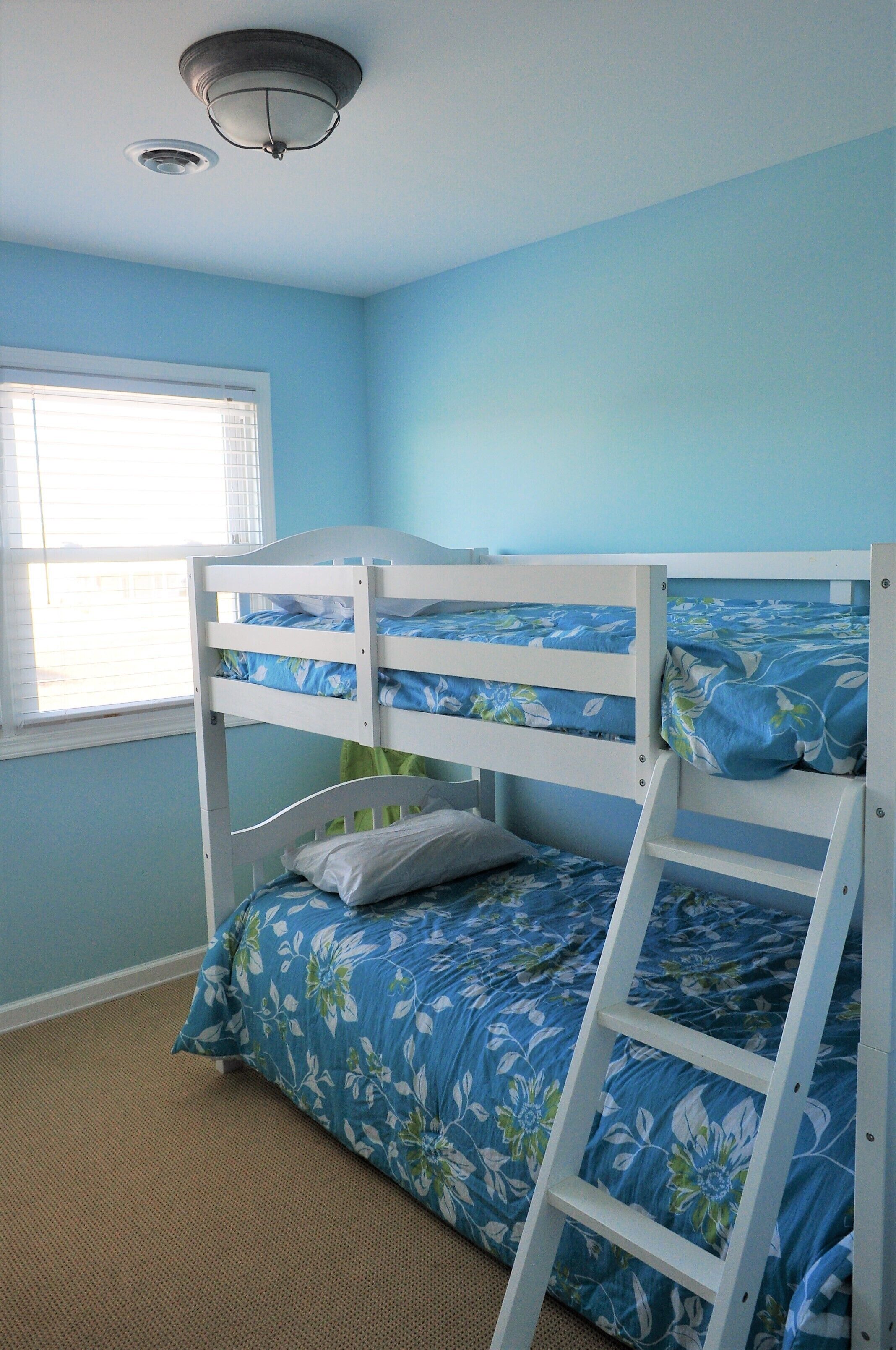 A bunk bed with floral bedding, a window with blinds, and a carpeted floor.