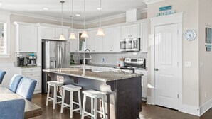 A modern kitchen with white cabinets, stainless steel appliances, a granite island, bar stools, pendant lighting, and a clock on the wall. - Experience ocean views at Panama Breeze, a spacious 6-bedroom retreat in Oak Island. (Oak Island)