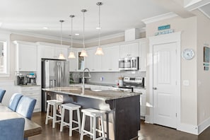 A modern kitchen with white cabinets, stainless steel appliances, a granite island, bar stools, pendant lighting, and a clock on the wall.