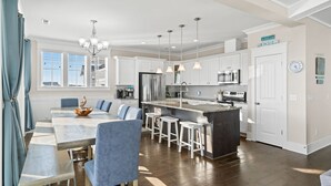 A modern kitchen with white cabinets, a central island with stools, a dining area with blue chairs, and a chandelier. - Experience ocean views at Panama Breeze, a spacious 6-bedroom retreat in Oak Island. (Oak Island)