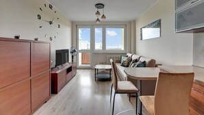 A modern living space with a wooden cabinet, a TV, a dining area with chairs, a sofa with cushions, and a large window with a city view. - RentPlanet - Apartament Przymorze (Gdańsk)