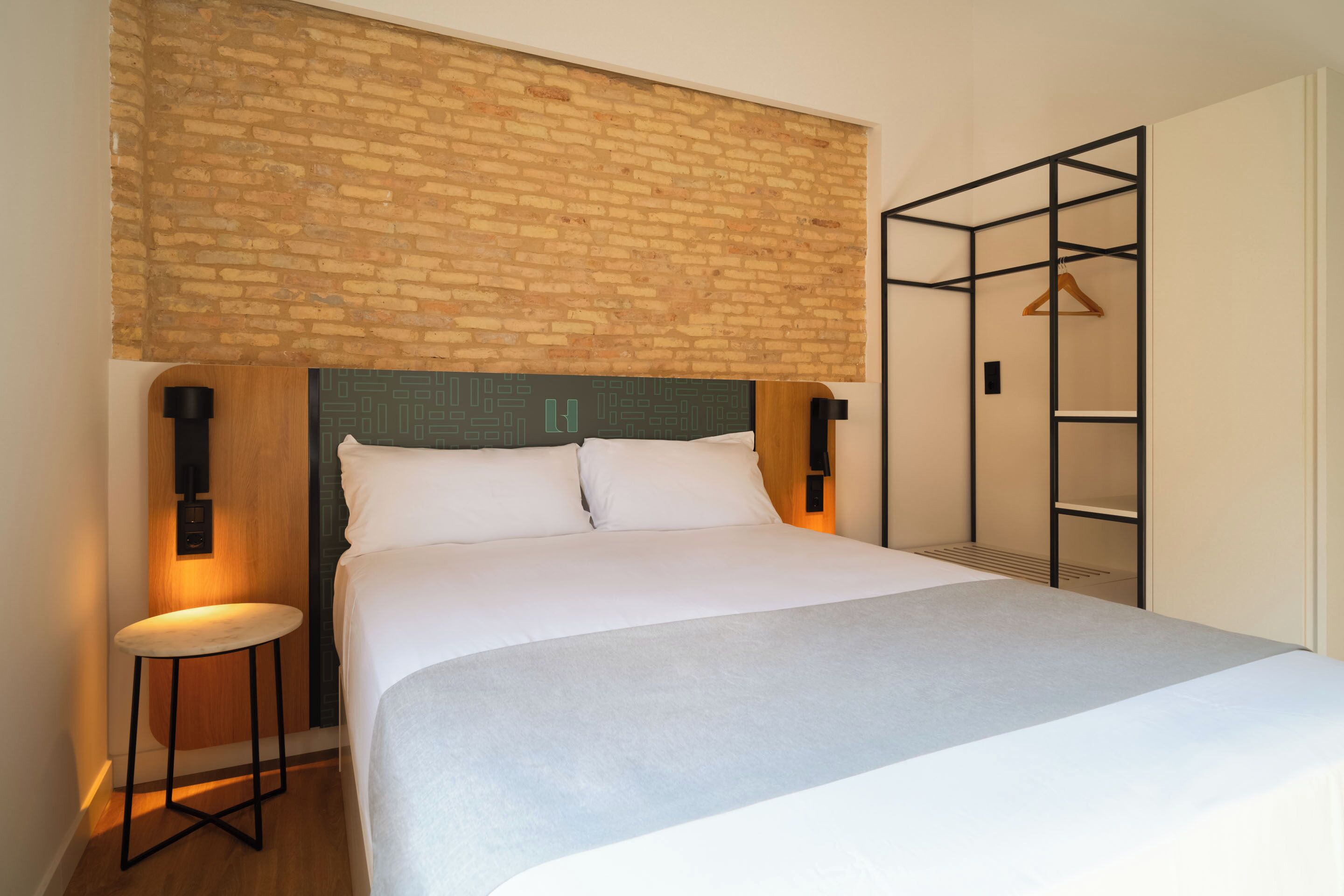 Une chambre à coucher moderne avec une tête de lit ornée d’un motif en briques, une table de chevet avec une lampe et une armoire intégrée dotée d’un portant à vêtements en bois.
