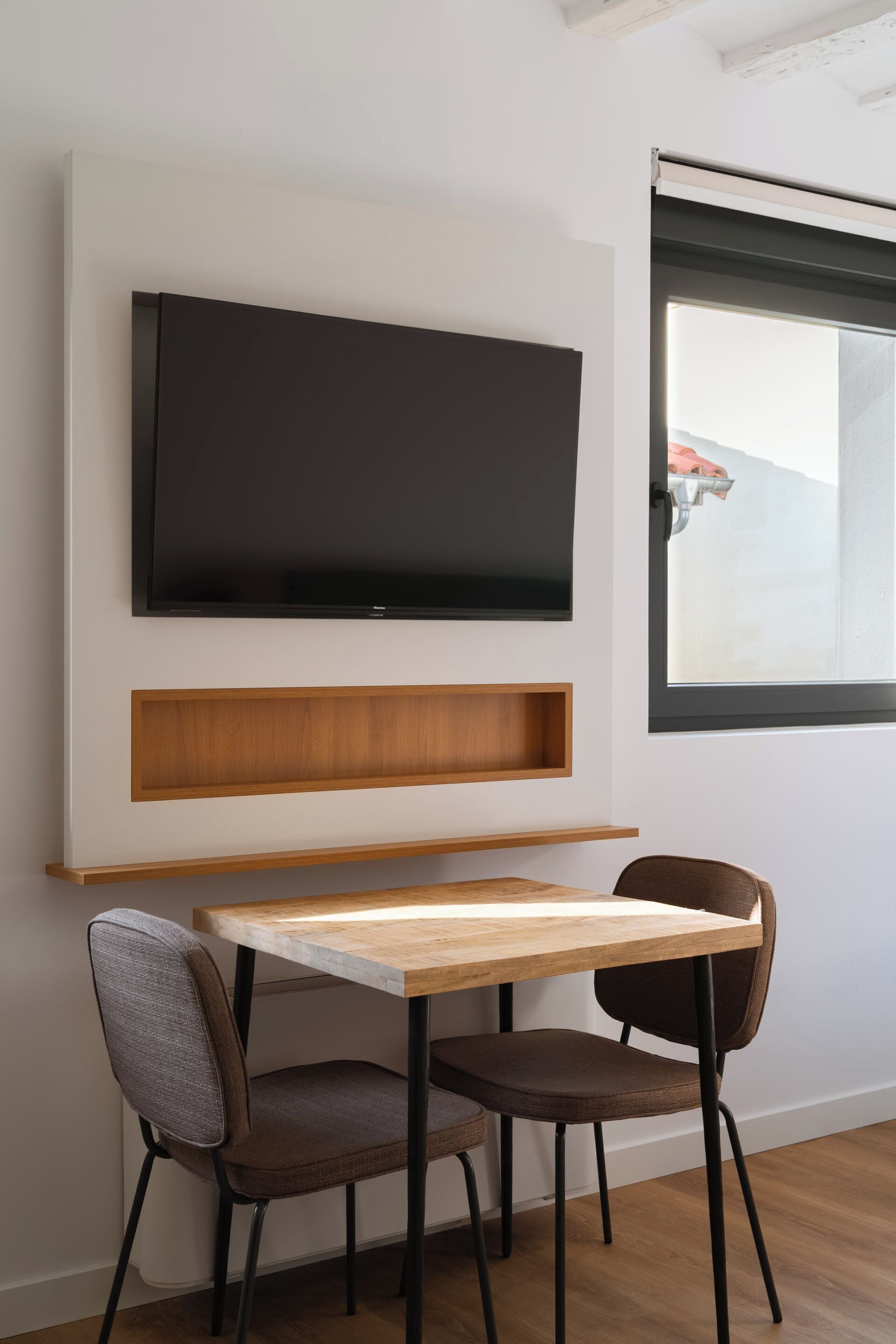 A modern dining area with a wooden table and two chairs, a flat-screen TV mounted on the wall, and a window with a view of a mountain.