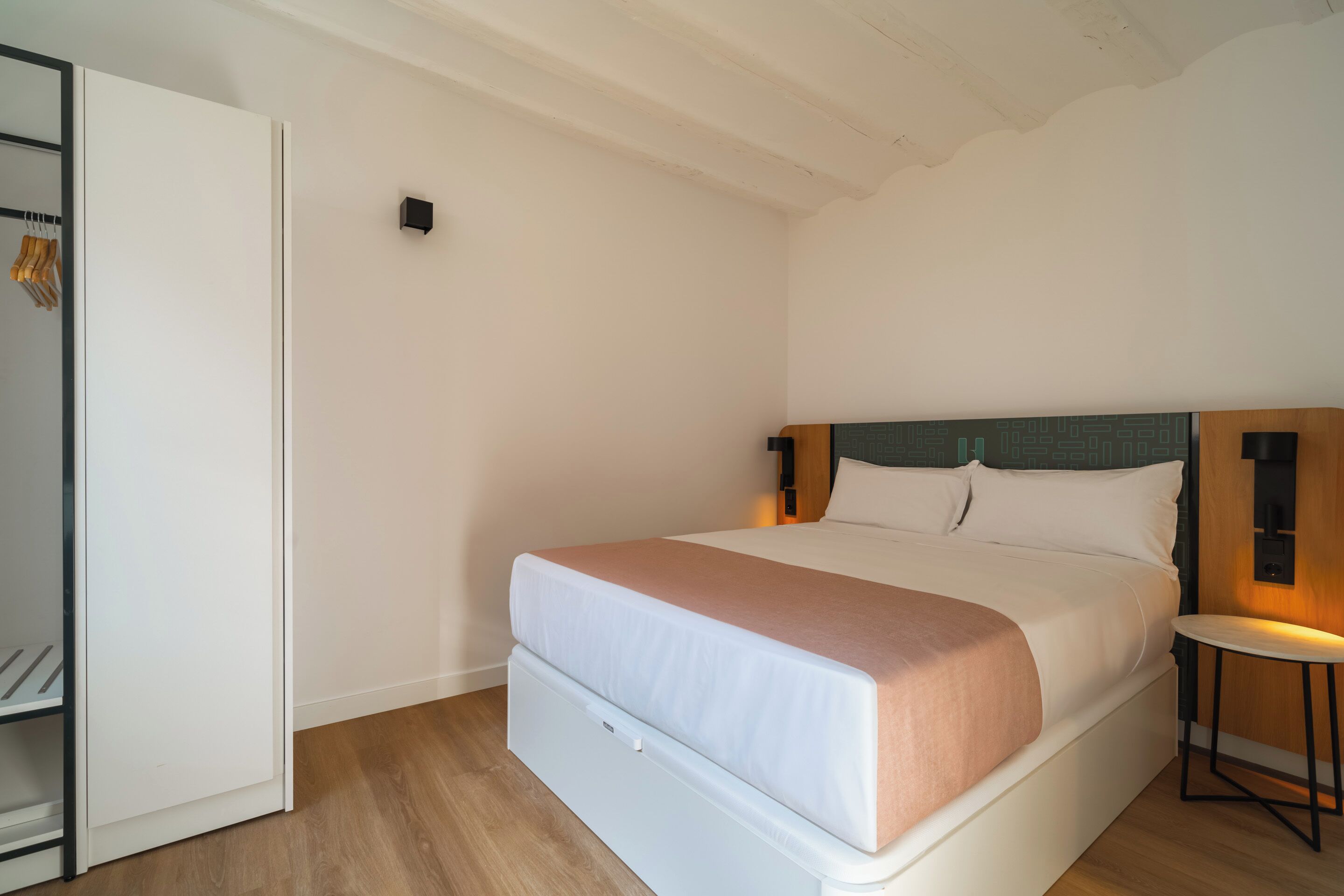 Un dormitorio con cama, mesita de noche, armario y un taburete pequeño.