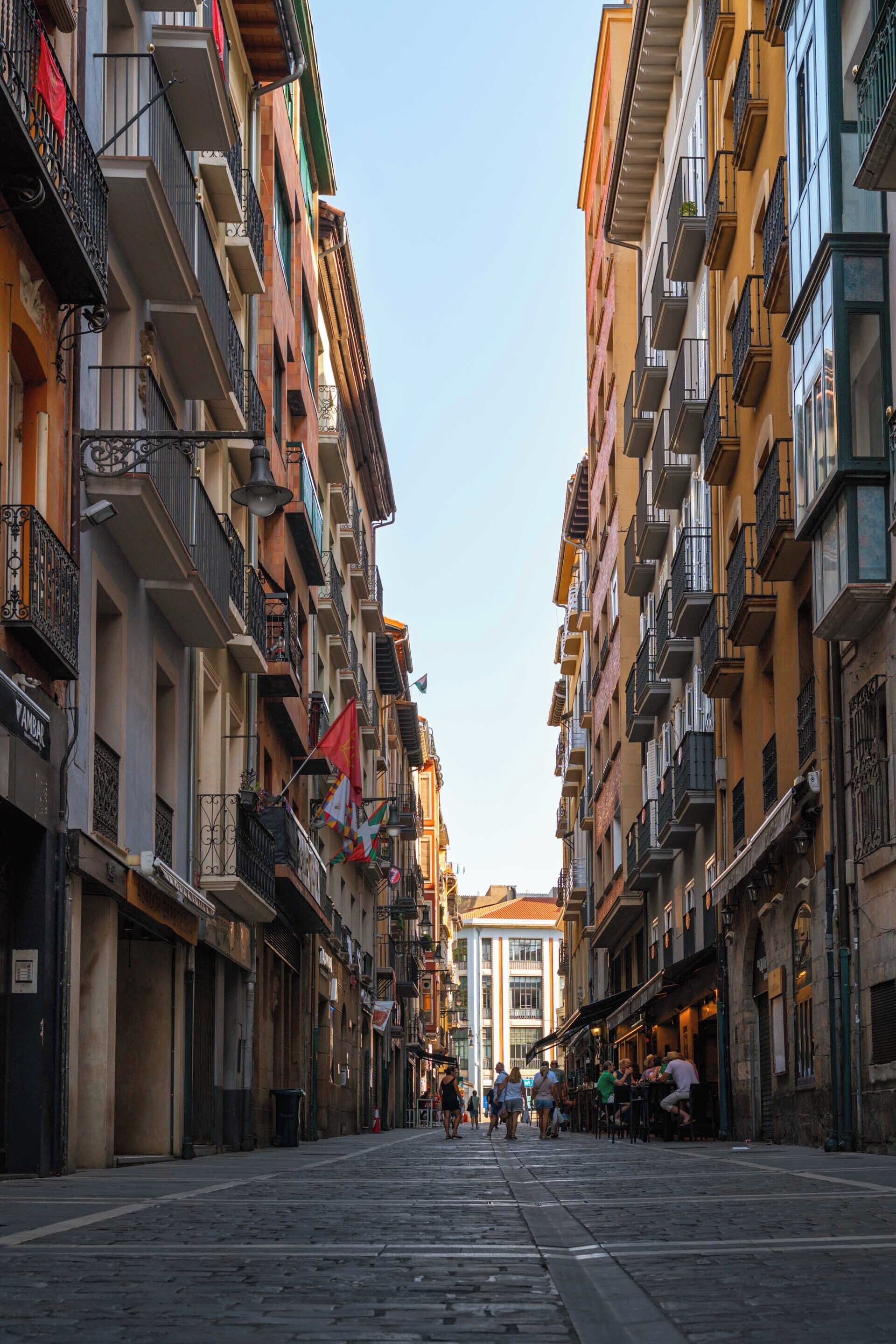 A narrow street flanked by multi-story buildings with balconies and outdoor seating areas.