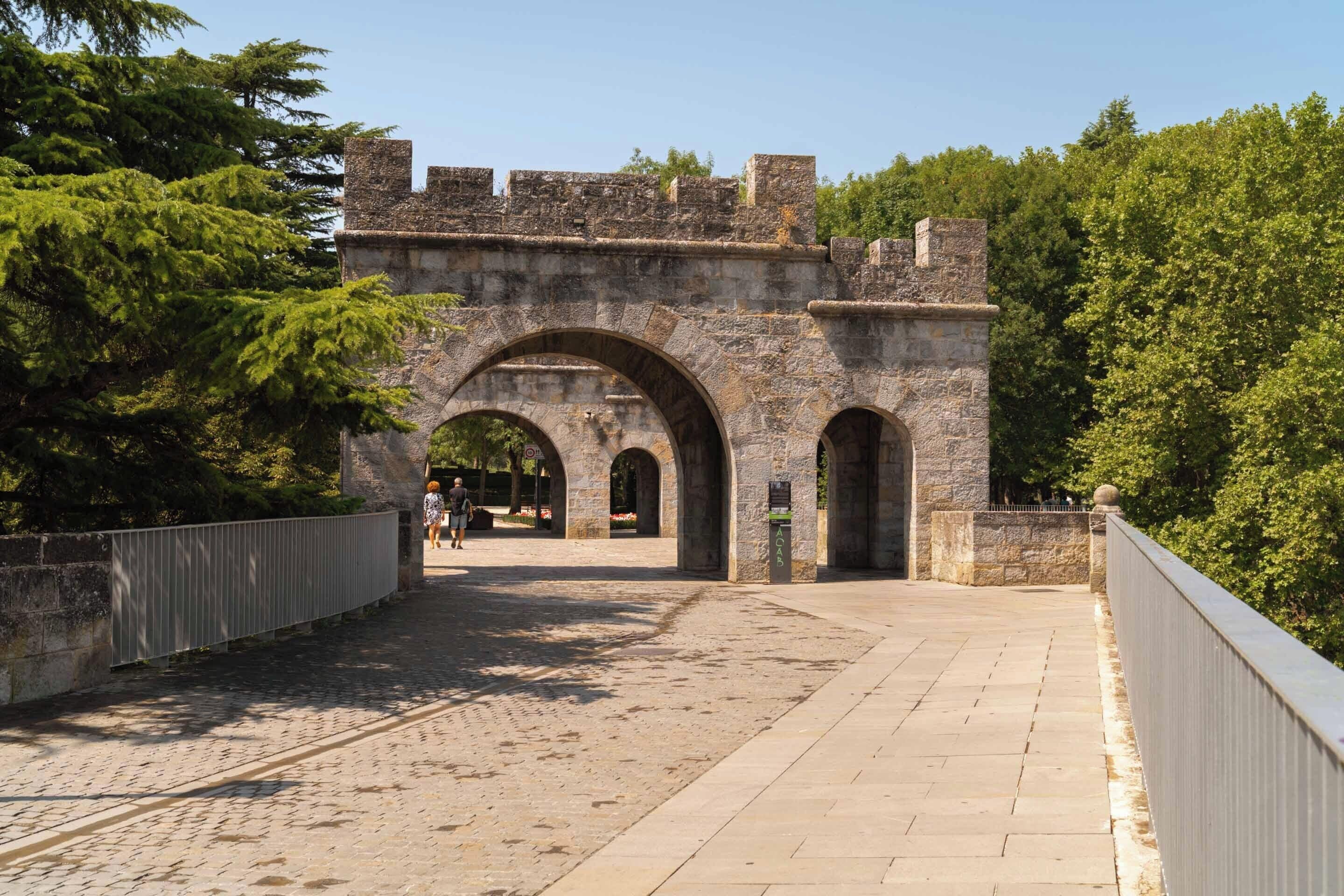 A stone bridge with arched passageways and a metal railing, surrounded by trees.