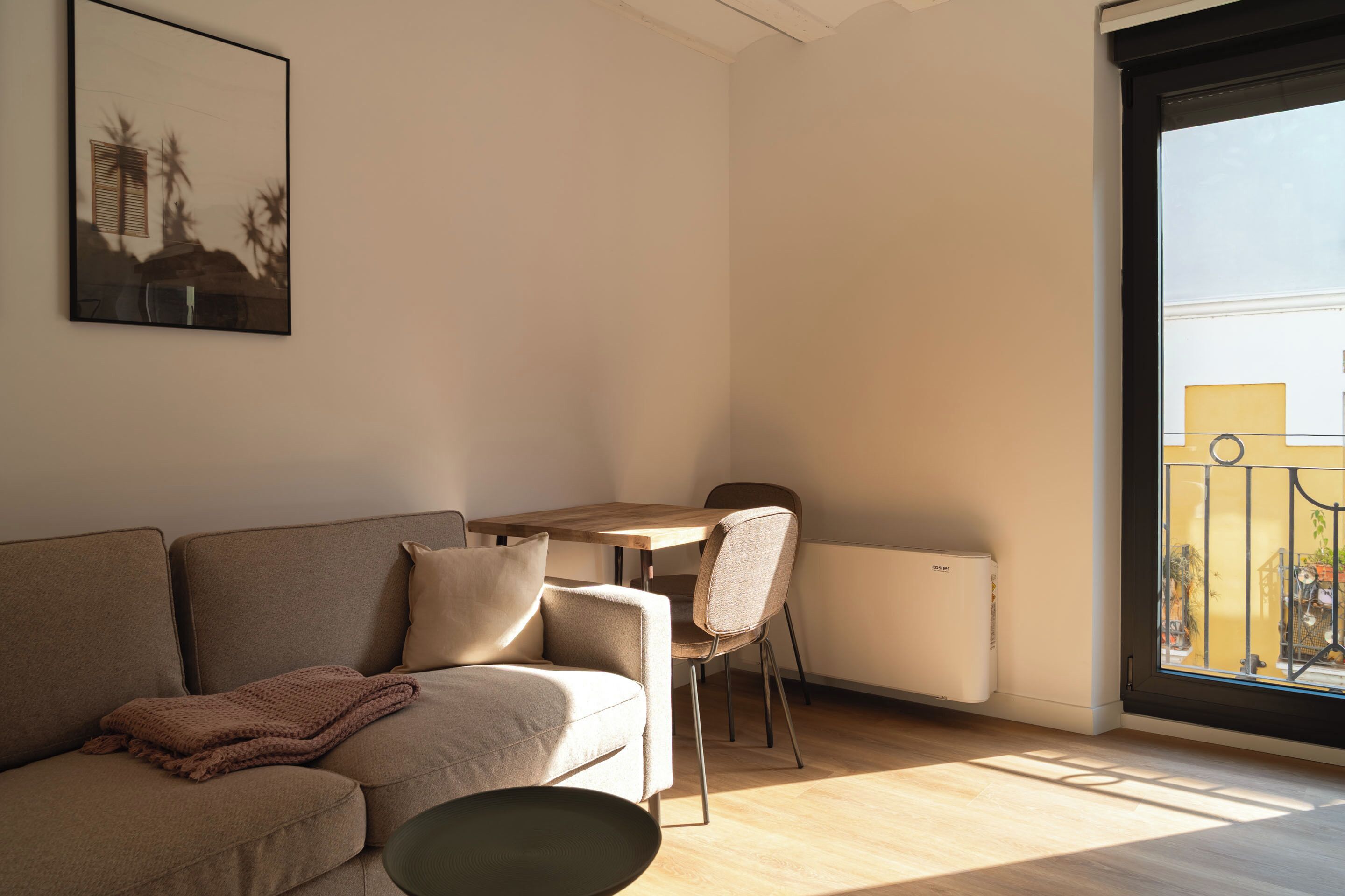 A modern living room with a sofa, a coffee table, and a dining table with chairs. There is a radiator and a window with a view of buildings.