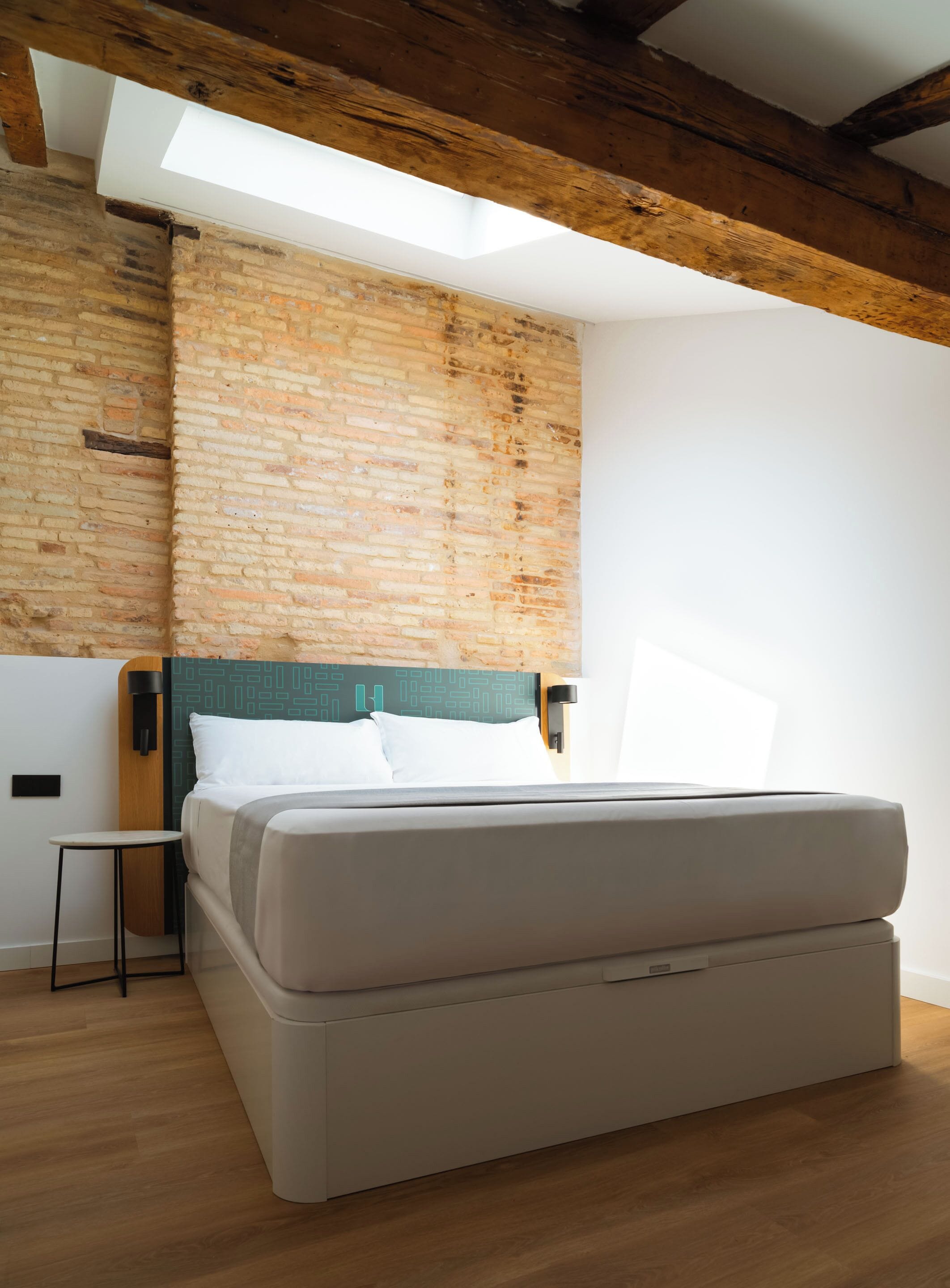 Un dormitorio moderno con una cama grande, una mesita de noche pequeña, cabecera con estampado geométrico y vigas de madera a la vista en el techo.