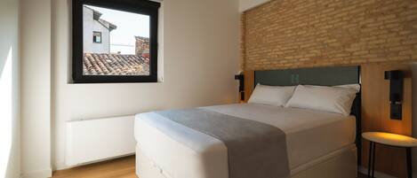Un dormitorio con una cama grande, una ventana con vistas a edificios, una pared de ladrillo y un taburete pequeño.
