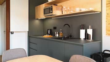 A compact kitchen with a microwave, sink, and modern appliances. There are wooden shelves above the counter holding dishes and glasses.