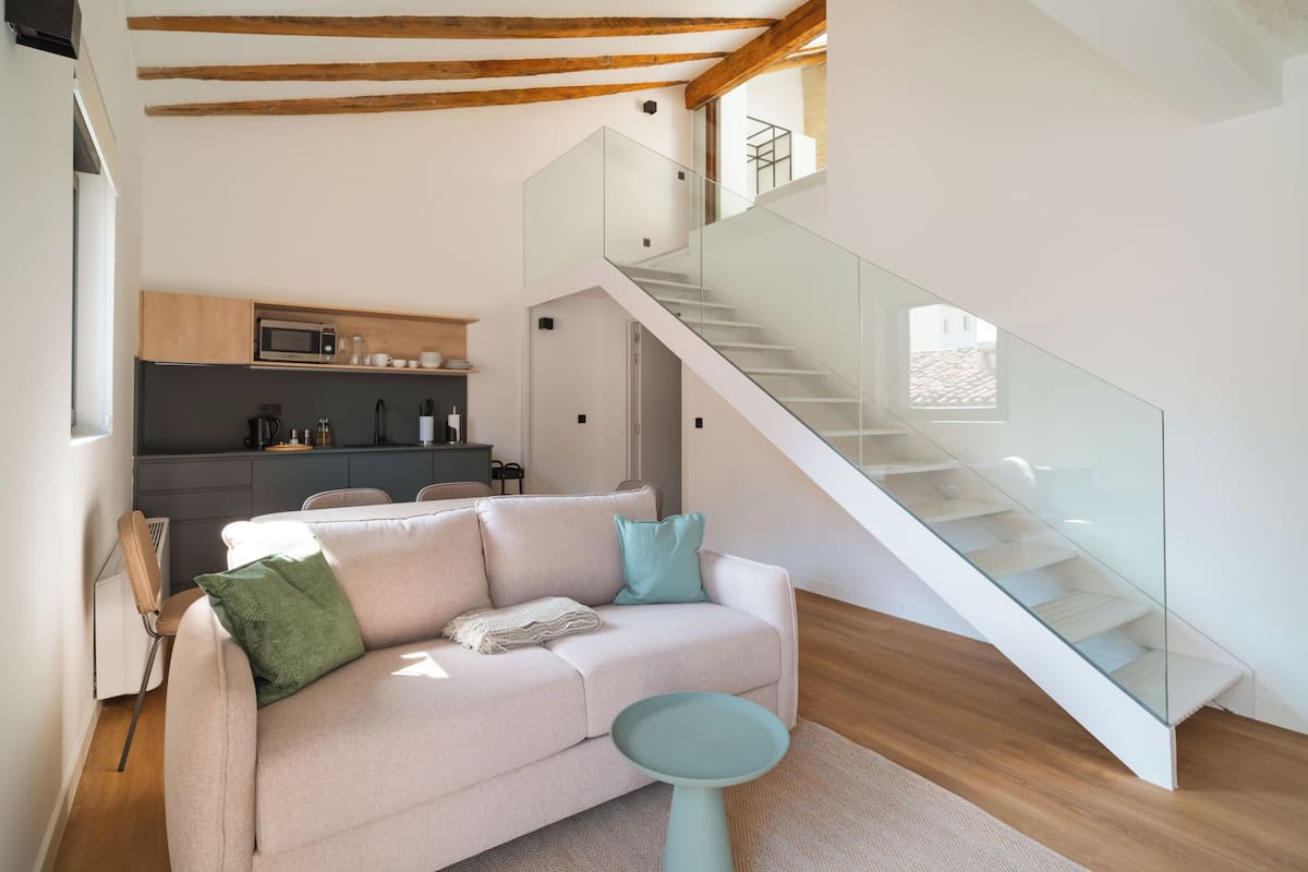 Uma sala de estar moderna com um sofá, uma escadaria de vidro e uma cozinha ao fundo.