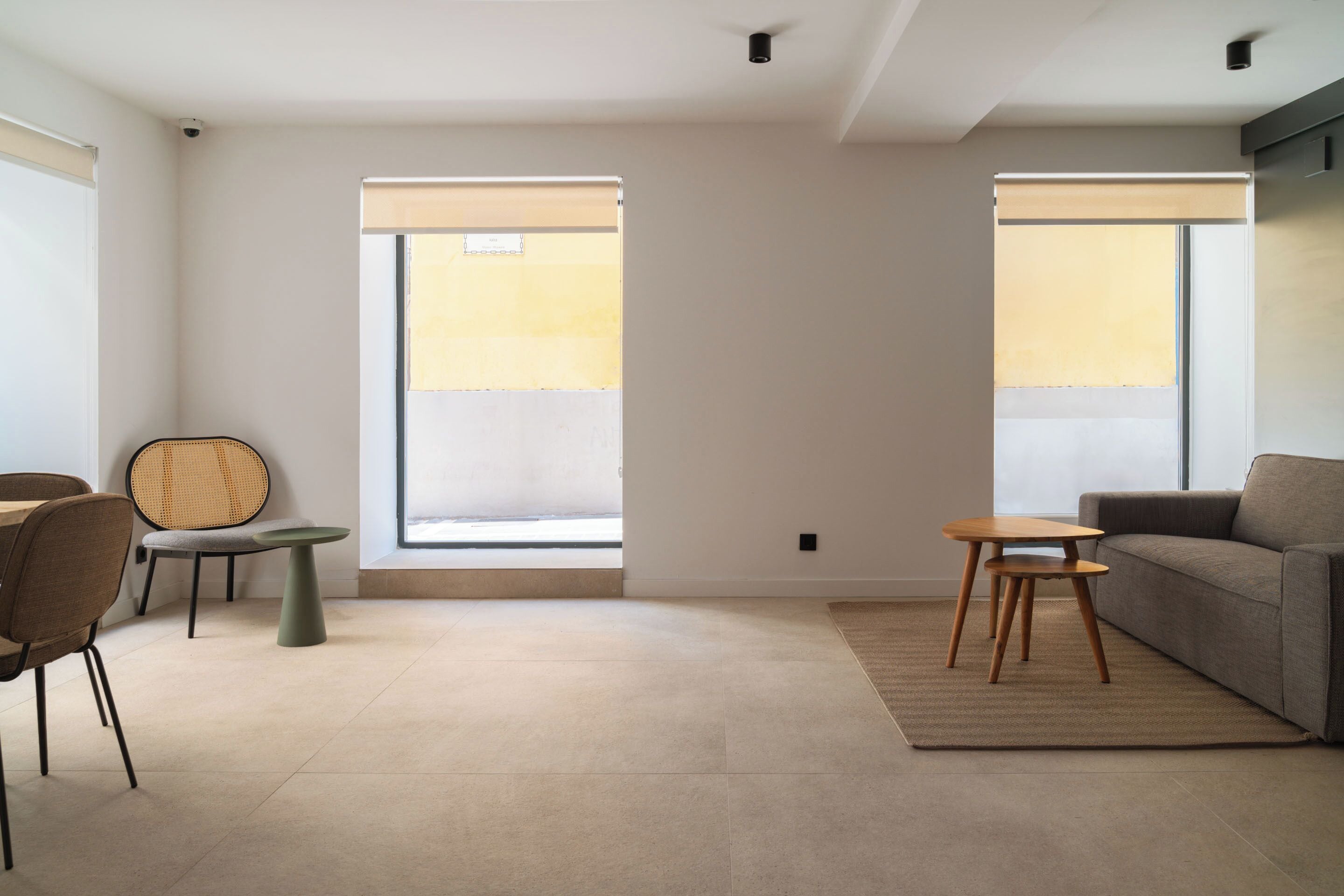 A modern living room with a sofa, a coffee table, and a chair. There are two windows with blinds, a ceiling light, and a wall-mounted security camera.
