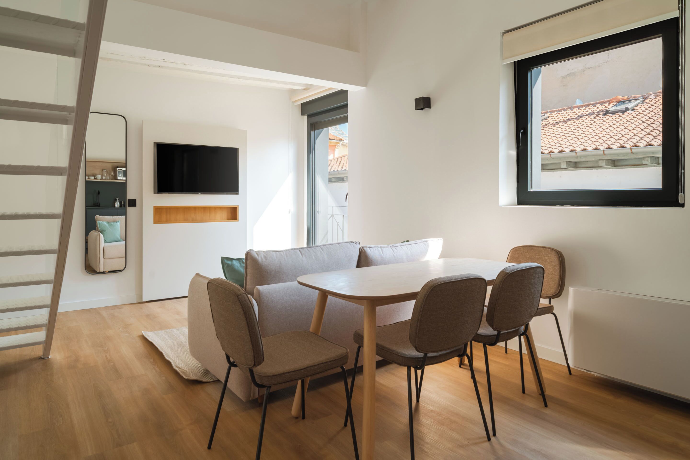 Una habitación moderna y bien iluminada con suelo de madera, una mesa blanca con cuatro sillas, un sofá, un televisor montado en la pared y una ventana con vistas a un tejado de baldosas.