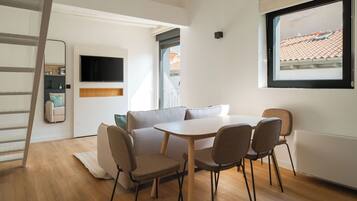 Una habitación moderna y bien iluminada con suelo de madera, una mesa blanca con cuatro sillas, un sofá, un televisor montado en la pared y una ventana con vistas a un tejado de baldosas.