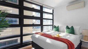 Ein modernes Hotelzimmer mit großem Fenster, einem Bett mit weißer Bettwäsche, grün-weißen Kissen und einer roten Tagesdecke.