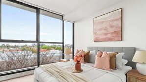 Ein Schlafzimmer mit großem Fenster, einem Bett mit vielen Kissen, einem Nachttisch mit Lampe und einem gerahmten Kunstwerk an der Wand.