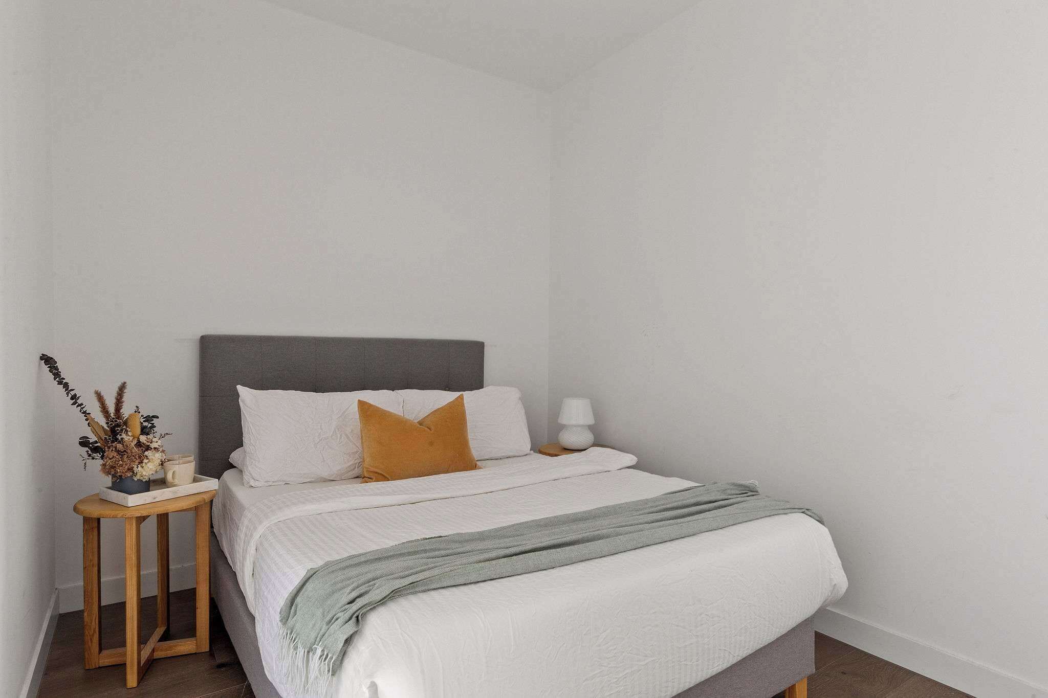 Un lit bien fait, avec une tête de lit grise, du linge de lit blanc et un plaid gris. Une petite table de chevet en bois, sur laquelle se trouve une lampe blanche et une plante décorative, est placée à côté du lit.