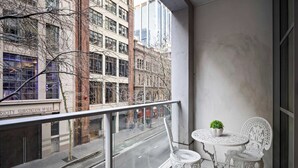 A balcony with a glass railing, two white chairs, a small table with a potted plant, and a view of a city street with buildings and a bicycle lane. - A Stylish Studio for 6, Next to Darling Harbour (Sydney)