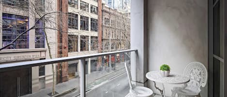 A balcony with a glass railing, two white chairs, a small table with a potted plant, and a view of a city street with buildings and a bicycle lane.