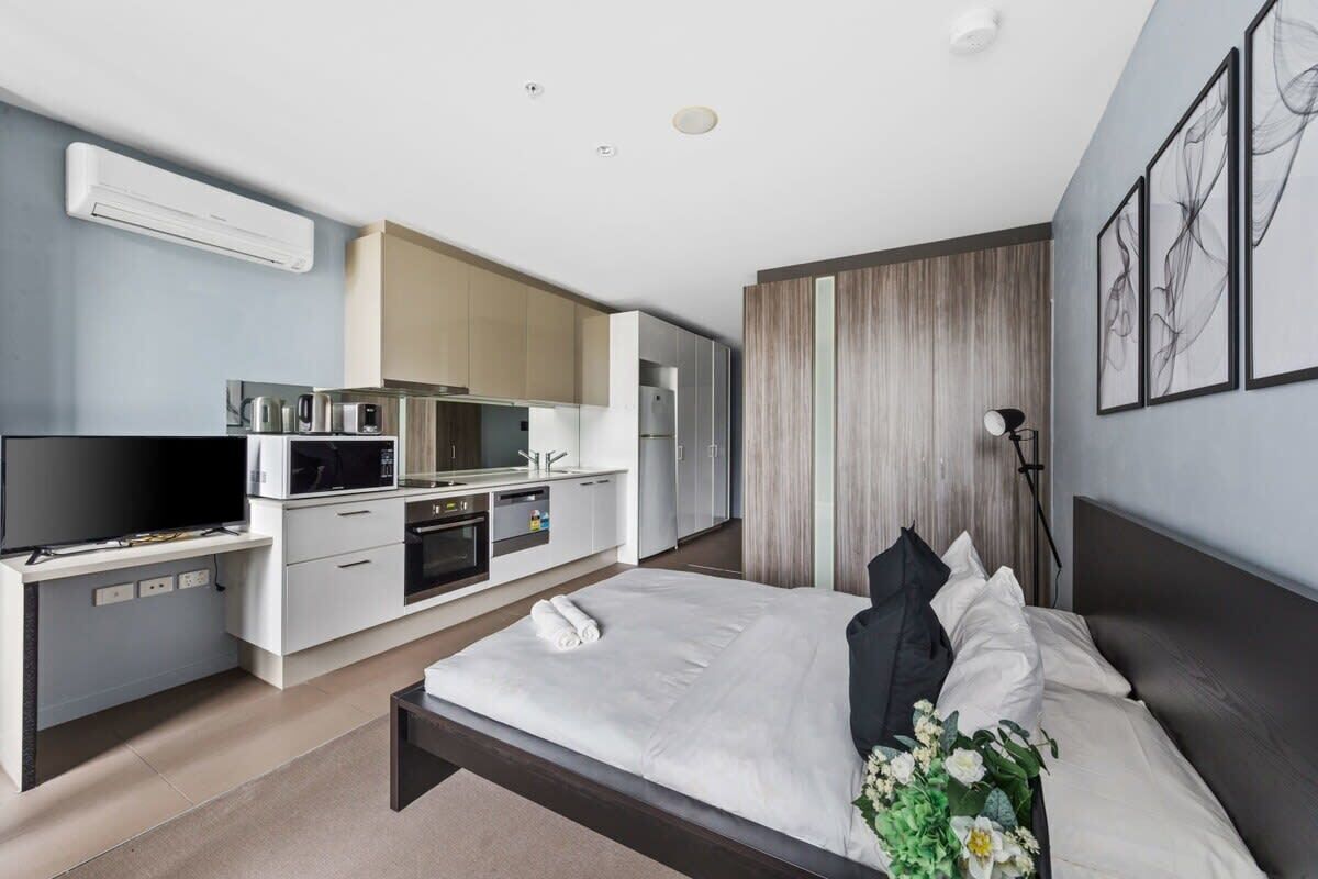 A modern hotel room with a large bed, a TV, a kitchenette, and a view of the city.