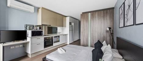 A modern hotel room with a large bed, a TV, a kitchenette, and a view of the city.
