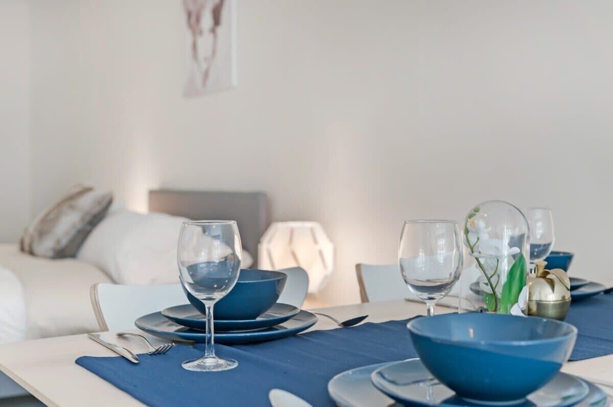 A dining table set with blue bowls, wine glasses, and cutlery, with a white sofa and a lamp in the background.