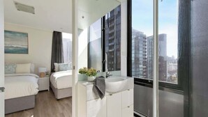 A modern hotel room with a large bed, a bedside table, a white bathroom with a sink, and a view of tall buildings.