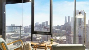 A modern living room with large windows, a city view, a table with a magazine, and a lamp.