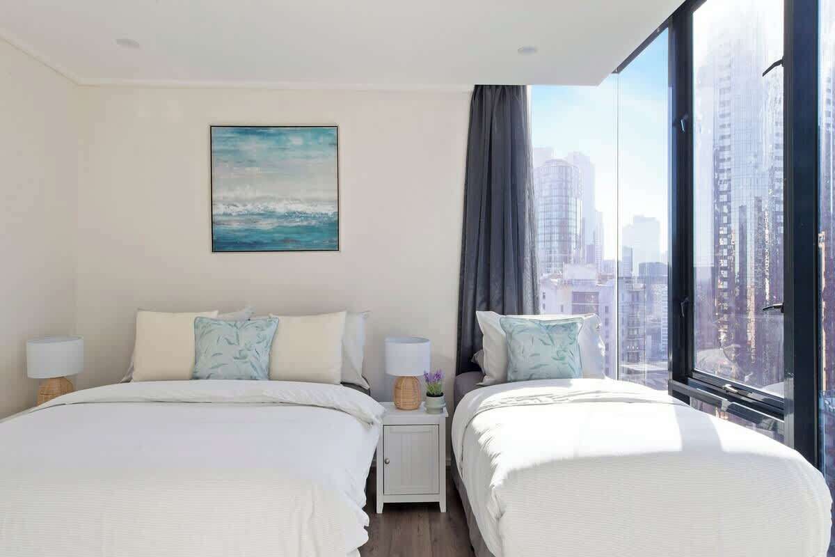 Habitación de hotel con dos camas, una mesita de noche con lámparas, un cuadro en la pared y un ventanal con vistas a la ciudad.