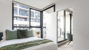 Ein modernes Schlafzimmer mit einem großen Bett, Blick auf eine Stadtlandschaft durch raumhohe Fenster und einem Balkon mit einem Sessel.