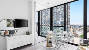 A modern living room with a large window, a flat-screen TV, a white sofa, and a dining area with a table and chairs. - Cozy & Central 2BR Stay, Near Cbd, Free Parking (Melbourne)