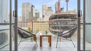 A balcony with two black wire chairs, a small glass table, and a view of a cityscape with tall buildings and a construction site. - Charming City-view Apt, Next to Darling Harbour (Sydney)