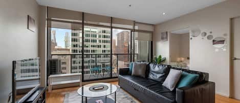 A modern living room with a leather sofa, a glass coffee table, and a view of city buildings through large windows.