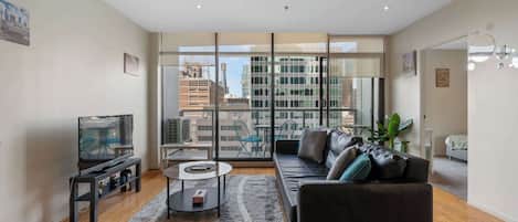 Una sala de estar moderna con sofá, mesa de centro y televisor. Amplios ventanales ofrecen vistas a la ciudad.