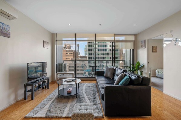 Ein modernes Wohnzimmer mit Sofa, Couchtisch und Fernseher. Große Fenster bieten Blick auf die Stadt.