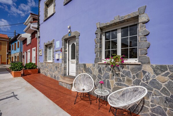 Un edificio con fachada de piedra, una puerta y una ventana con marco de piedra. Hay dos sillas de mimbre blanco y una mesita con un jarrón de flores.