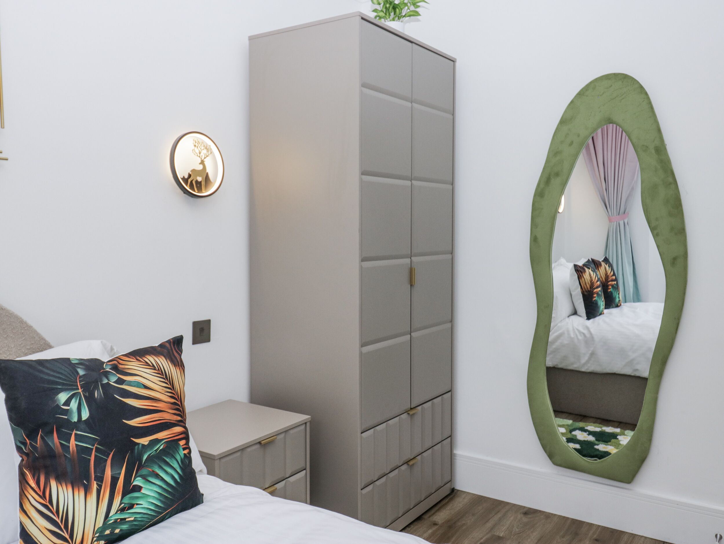 A bedroom with a bed, a grey wardrobe, a small bedside table, a wall-mounted mirror, and a wall clock.