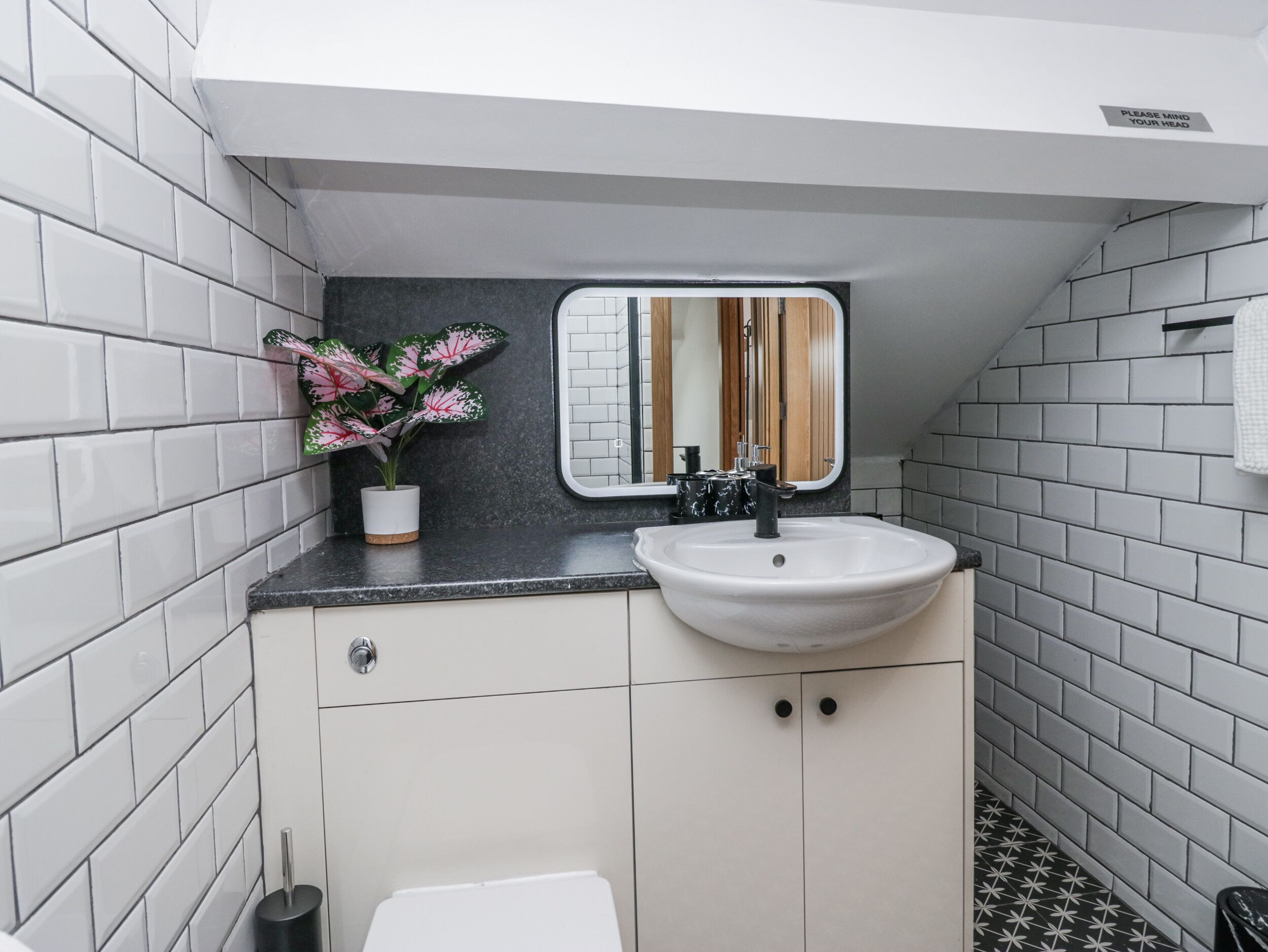 A bathroom with white tiled walls, a black countertop, a white sink, a mirror, and a potted plant.