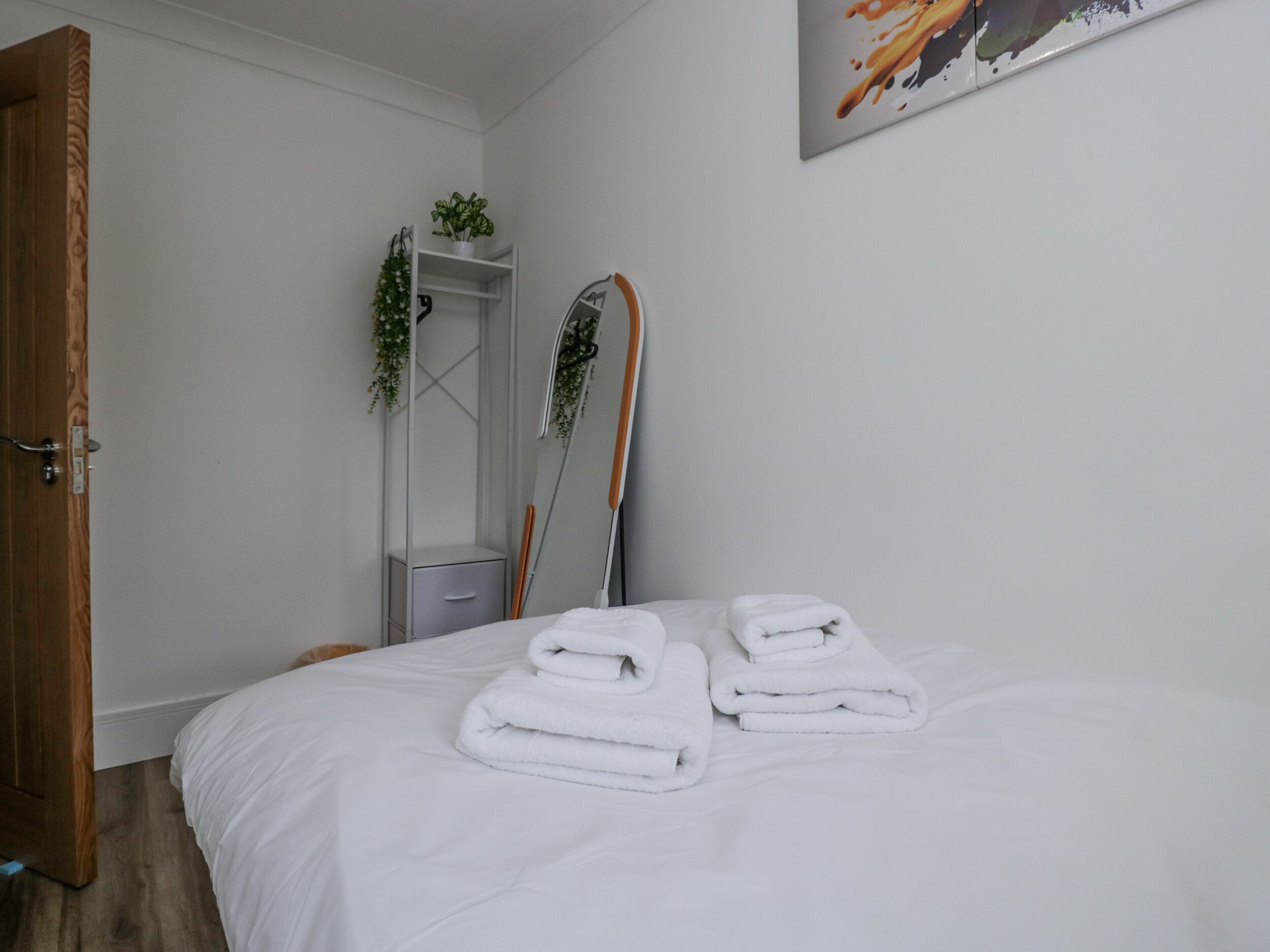 A hotel room with a bed, a door, a mirror, a towel rack, and a painting on the wall.