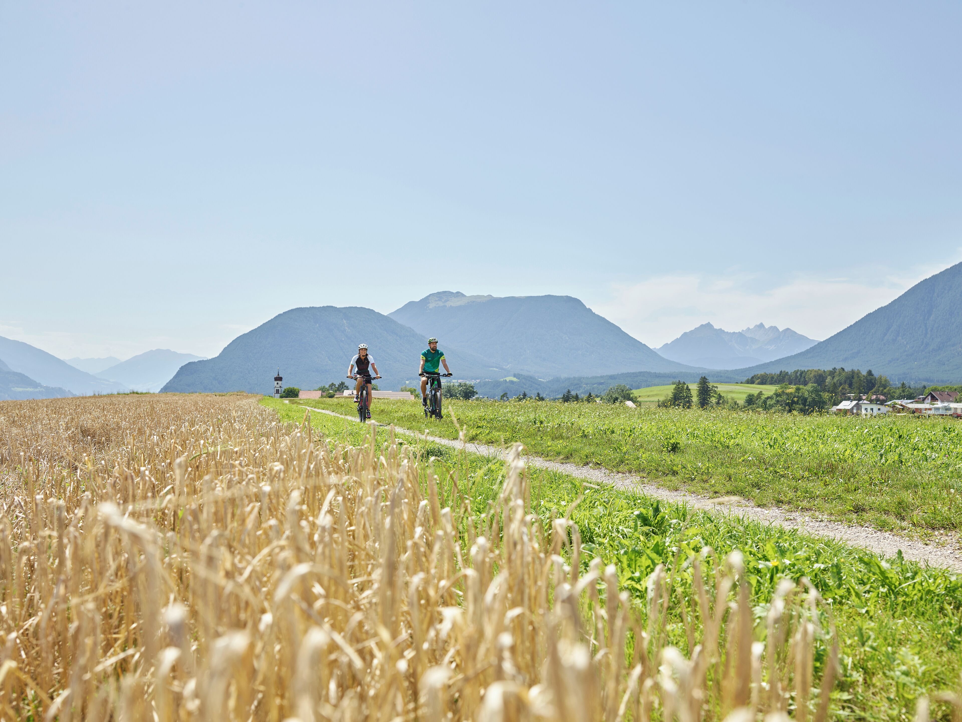 A rural landscape with a path, two cyclists, and mountains in the background.