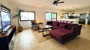 Un salon moderne avec un canapé, un fauteuil et un téléviseur, une cuisine avec des tabourets de bar et un coin repas avec une table et des chaises.