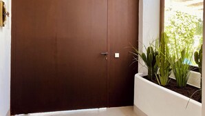 A large brown door with a handle, a white wall, and a window with a view of greenery outside. - Unique front line golf villa in Marbella (Marbella)